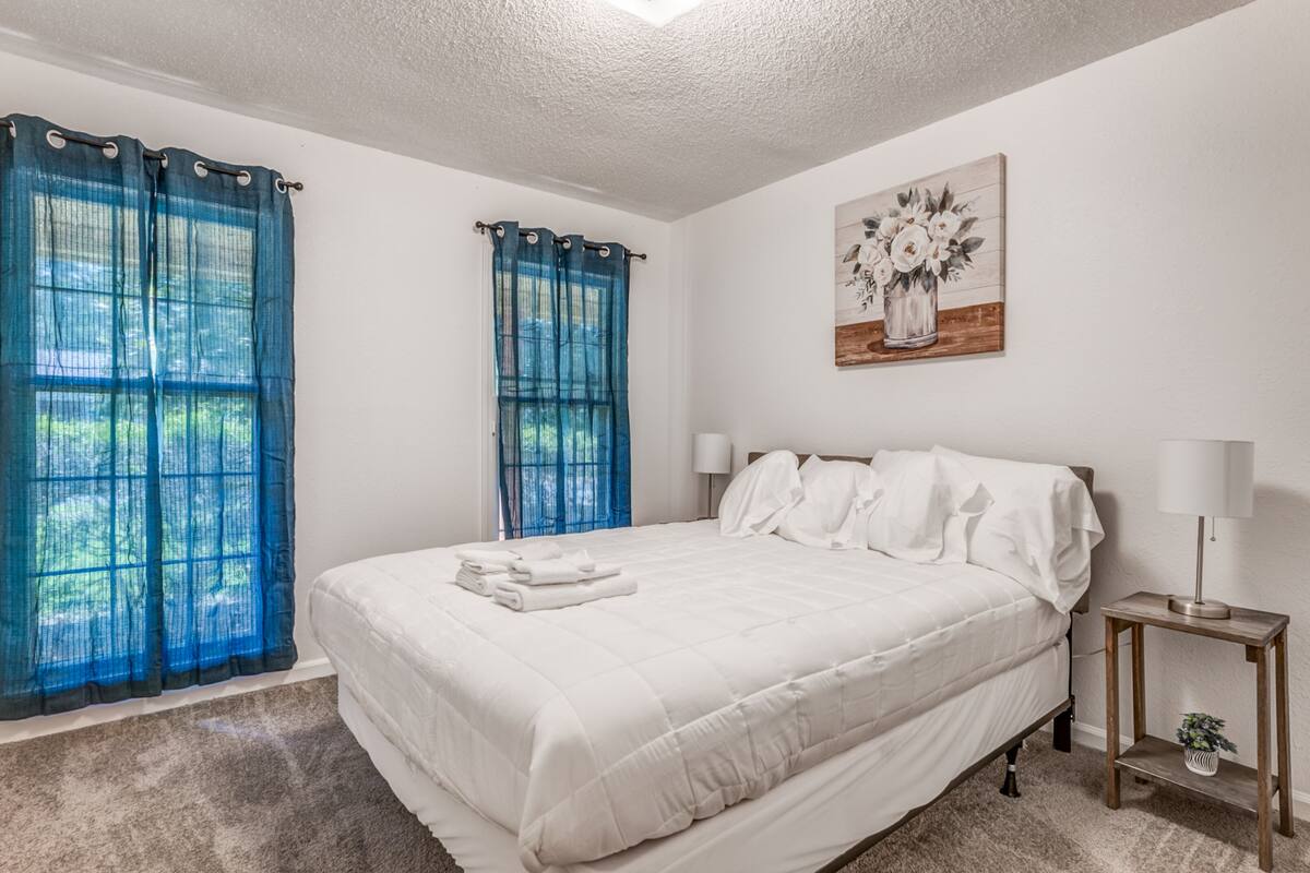 A bedroom with a large bed, two bedside tables, a window with blue curtains, and a painting on the wall.