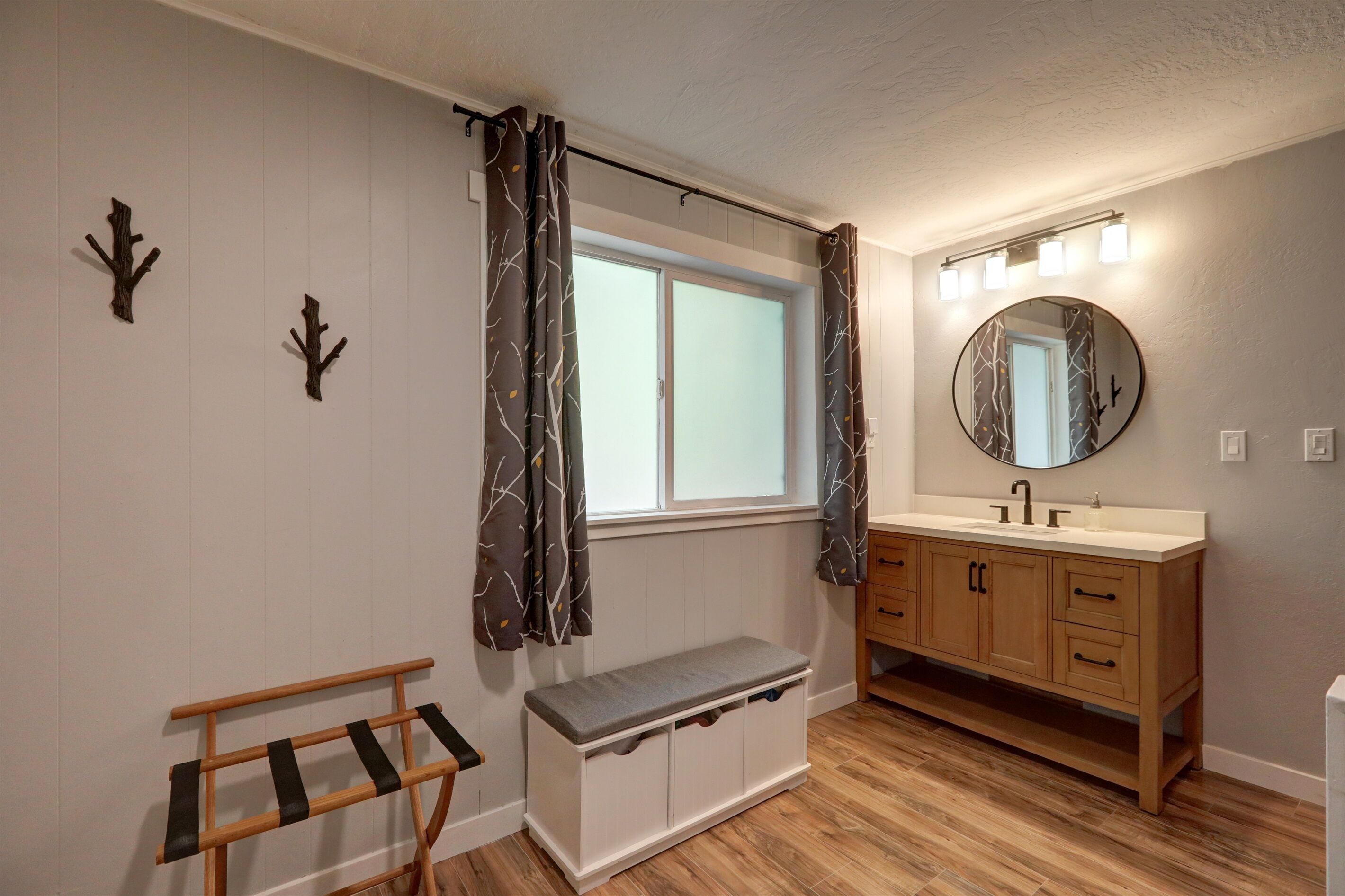 A bathroom with a wooden vanity, a round mirror with lights, a bench, and a window with curtains.