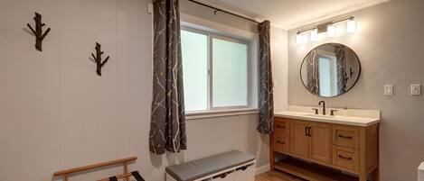A bathroom with a wooden vanity, a round mirror with lights, a bench, and a window with curtains.