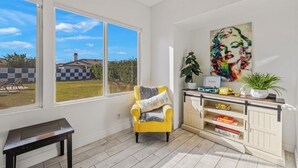 A room with a yellow armchair, a large window, a cabinet with decorative items, and a painting on the wall.