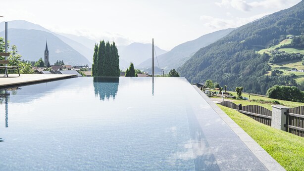 A clear view of a swimming pool with a mountain backdrop, a church spire, and a well-maintained lawn area. - Hotel Lahnerhof (Vipiteno)