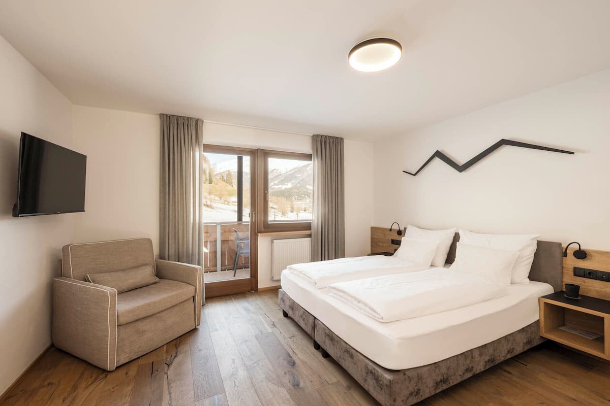 Quarto de hotel com cama, uma cadeira, televisão e vista para uma paisagem nevada.