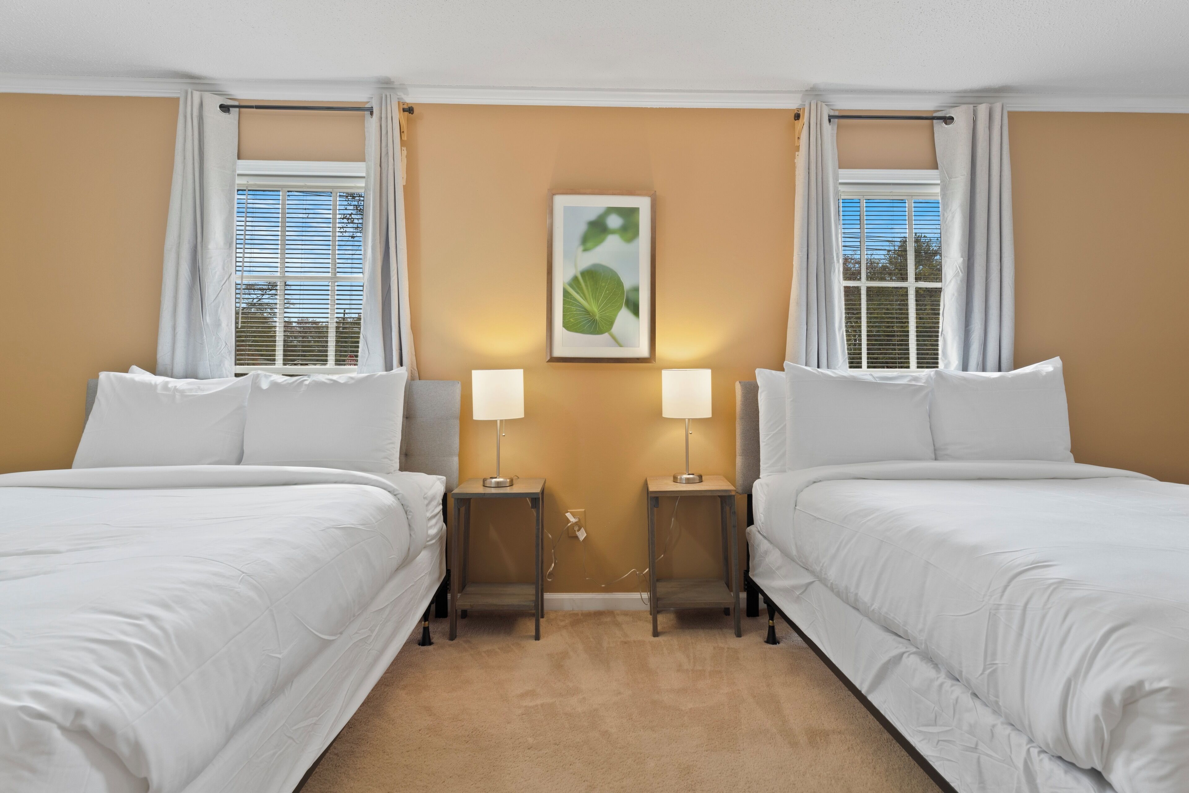 Ett hotellrum med två separata sängar, ett beigefärgat golv, ett inramat konstverk med ett lövmotiv på väggen och två sänglampor.