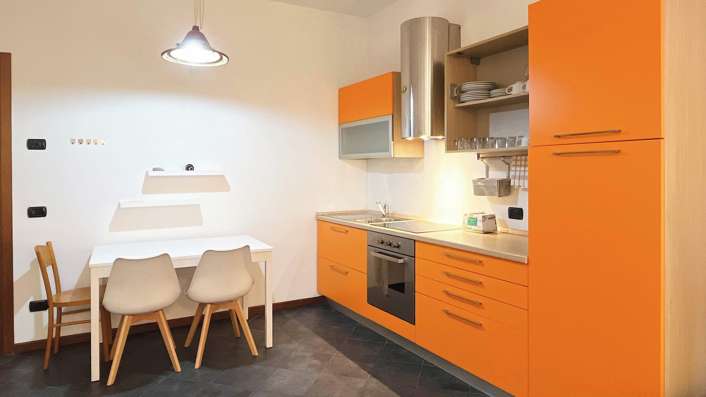 Een compacte keuken met een witte eettafel en stoelen, een inbouwoven en oranje keukenkastjes.