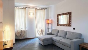 A modern living room with a grey sectional sofa, a floor lamp, a mirror with an ornate frame, and a window with white curtains. - Casa Marta - Affitti Brevi Italia (Lecco)
