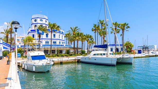 A marina with boats docked, a building with blue and white architecture, and palm trees. - Exclusive penthouse with 2 bedrooms and sea views  (Estepona)