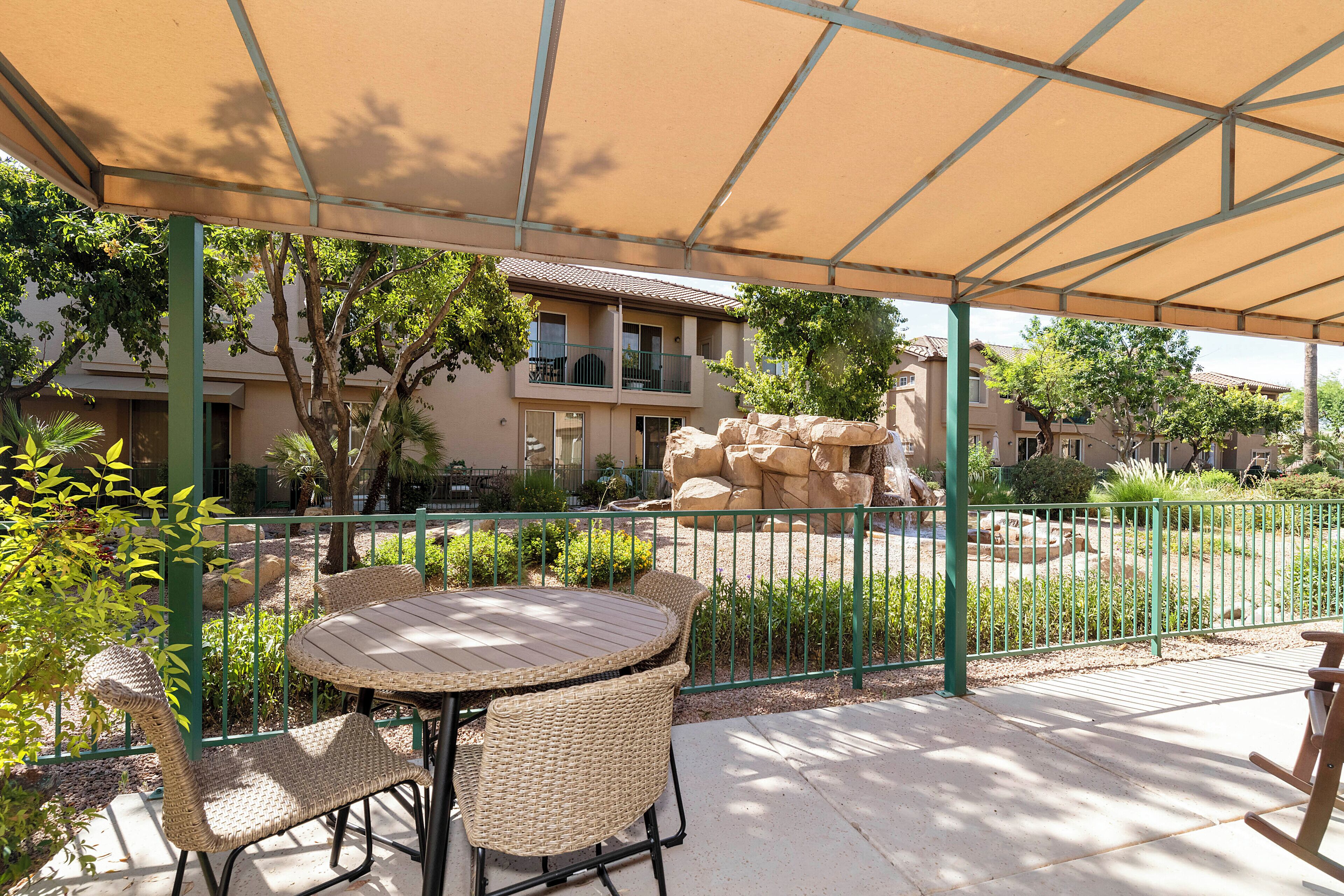 Ein Terrassenbereich im Freien mit einem runden Tisch und Korbstühlen, umgeben von einem grünen Metallzaun und einem beigen Sonnensegel.