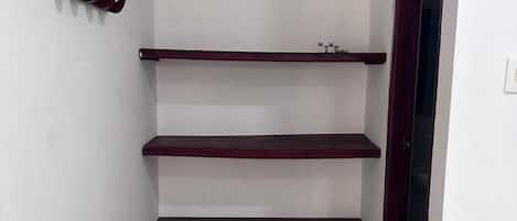 A small, narrow room with a white wall and a tiled floor. There are three wooden shelves mounted on the wall, and a door on the right side.