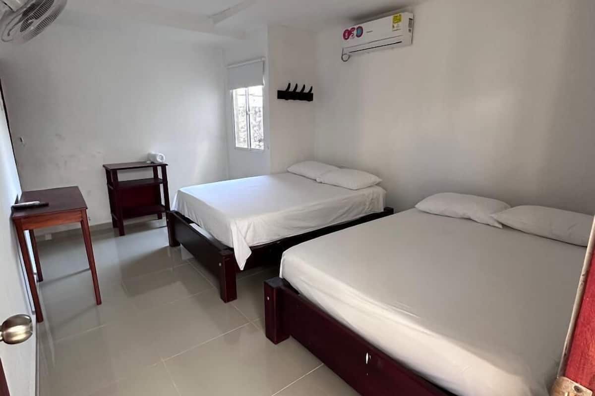 A room with two beds, a small table, a fan, and a window.