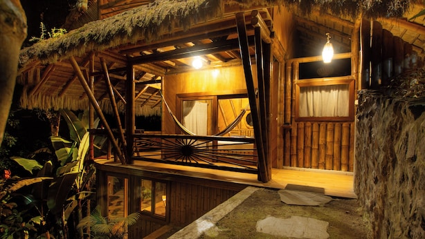 A rustic wooden cabin with a thatched roof, a hammock, and a small balcony overlooking a garden.