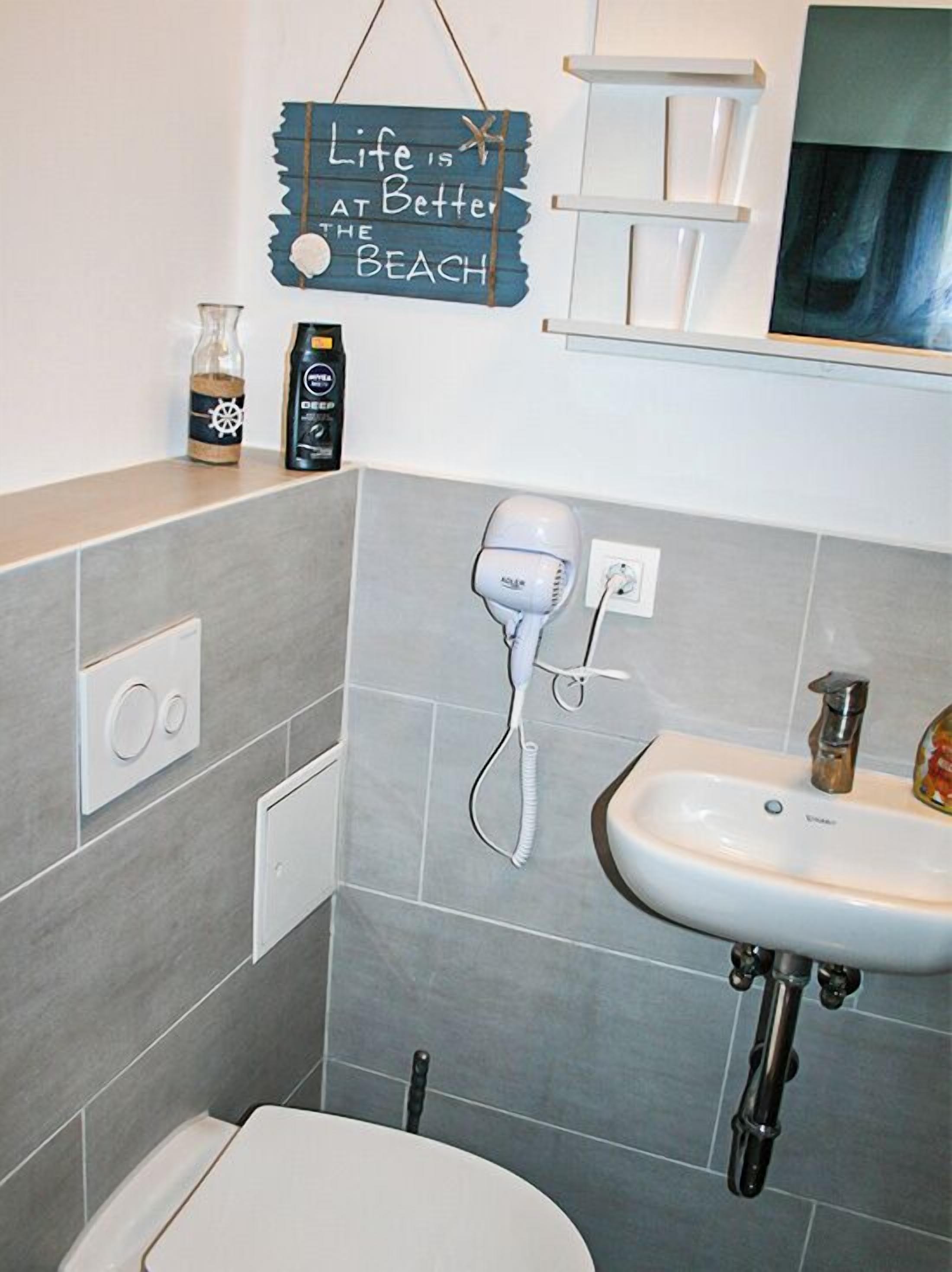 Casa de banho com lavatório branco, secador de cabelo, frasco de champô e placa com o dizeres 'A vida é melhor na praia'.