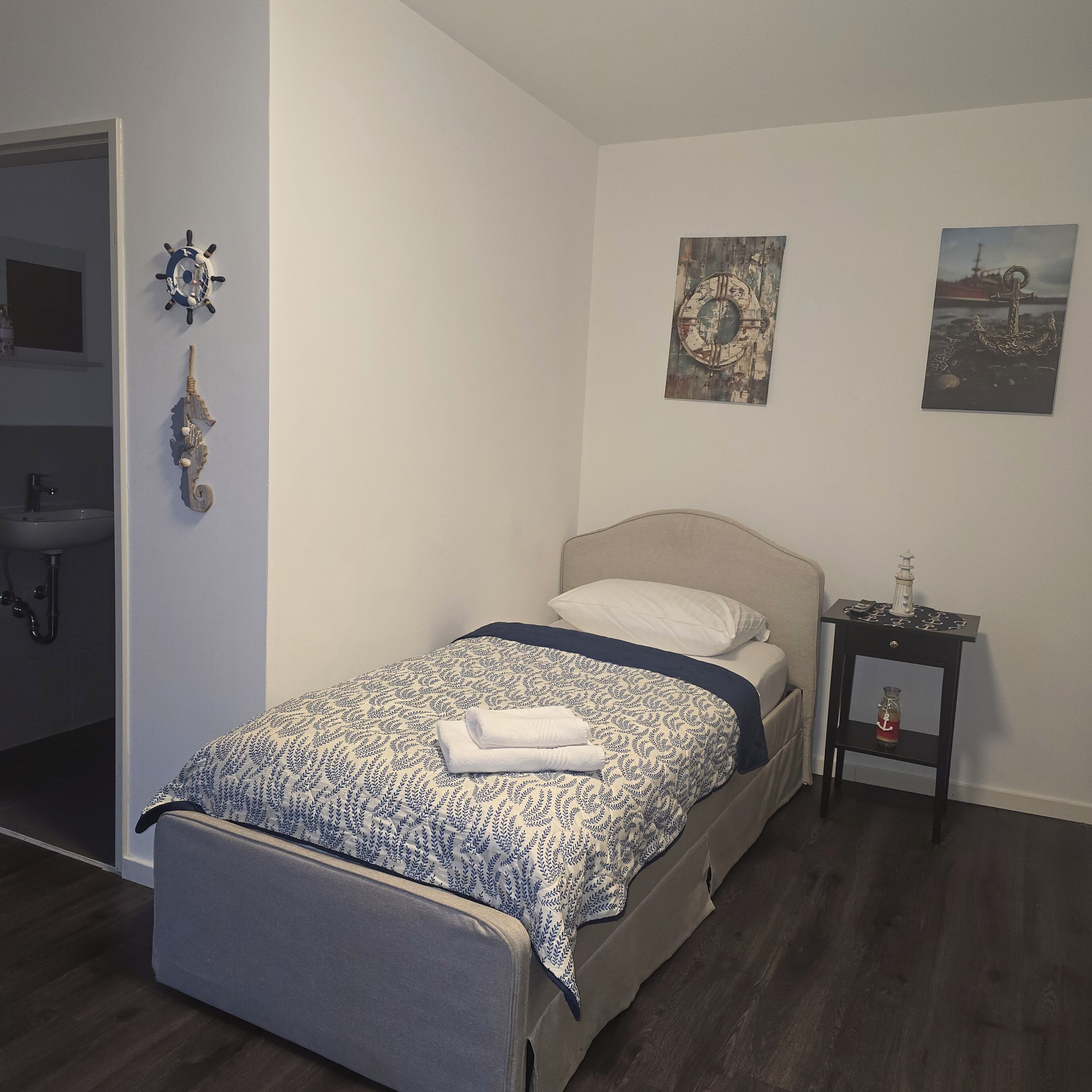 A neatly arranged bedroom with a bed, a bedside table with a bottle, and a wall-mounted ship's wheel.