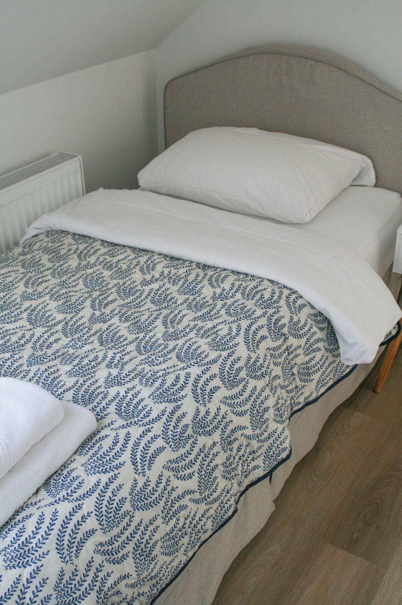 A bed with a white and blue patterned bedspread, a grey headboard, and a white pillow.