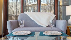 A modern living room with a grey sofa, two glass wine glasses on a glass table, and a view of trees through large windows. - Cozy and serene fishing cabin, Family Traditions,  private dock and lake views. (Gainesville)