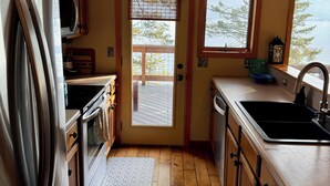 A kitchen with wooden cabinets, a stainless steel refrigerator, a microwave, an oven, a sink, and a window overlooking a deck.