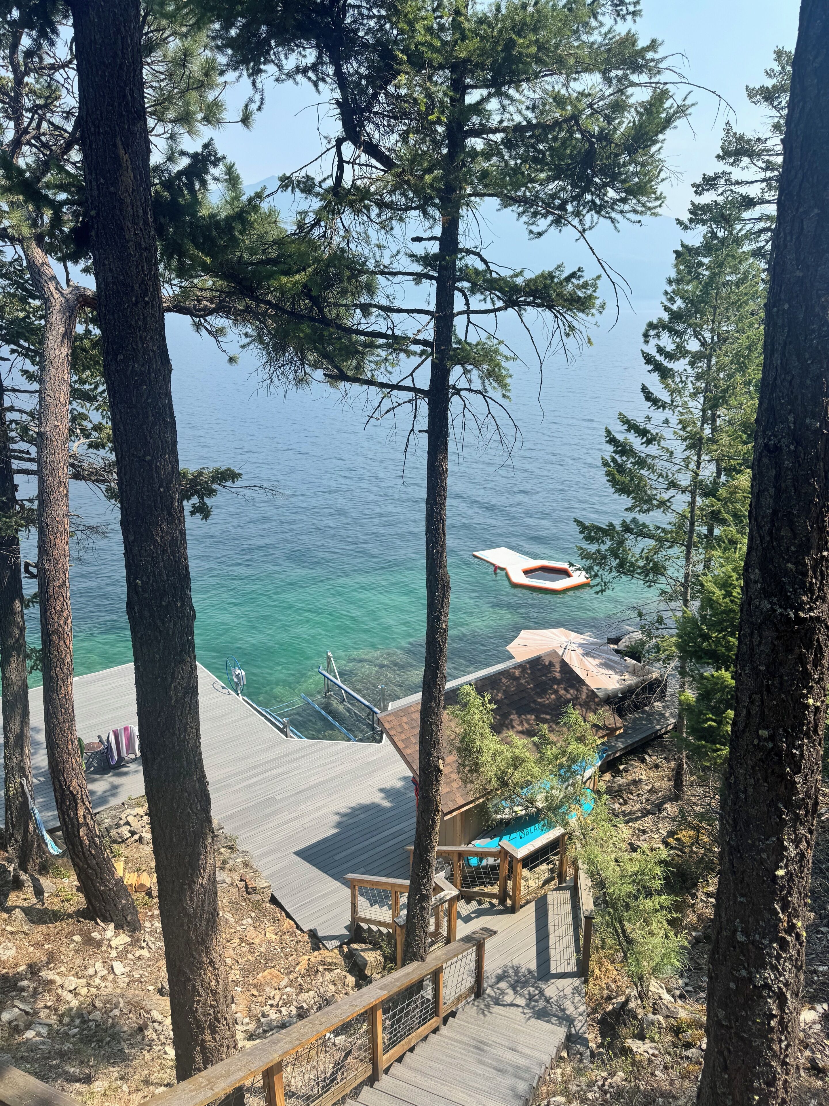 A wooden deck with a railing, leading to a dock over water, surrounded by trees.