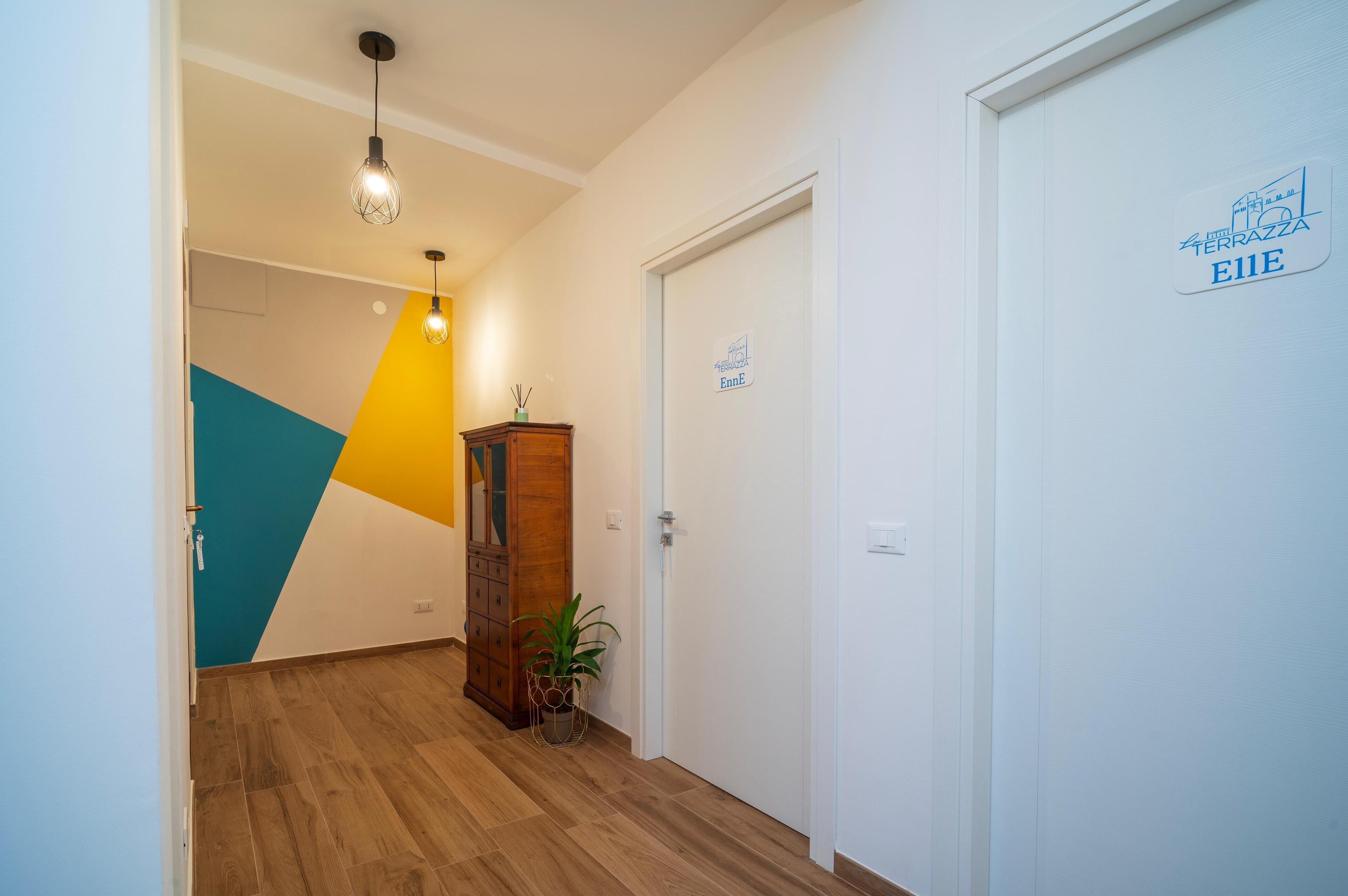 Un pasillo de hotel con piso de madera, diseño geométrico en la pared, un mueble de madera, una planta en maceta y una puerta con un letrero.