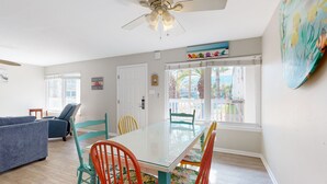 A dining area with a glass table and colorful chairs, a ceiling fan, and a view of the outdoors through large windows. - Beach Access Across the Street, Shared Heated Pool (Corpus Christi)