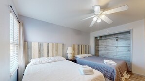 A bedroom with two beds, a ceiling fan, a window with blinds, and a wooden headboard. - Beach Across the Street, Shared Heated Pool, Pet Friendly (Corpus Christi)