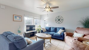 A living room with a ceiling fan, a television, a coffee table, sofas, and a wall clock. - Beach Across the Street, Shared Heated Pool, Pet Friendly (Corpus Christi)
