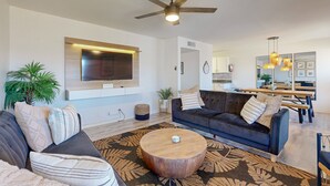 A modern living room with a large flat-screen TV, a dark-colored sofa, a round wooden coffee table, and a ceiling fan.