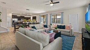 A spacious living room with a large sofa, a coffee table, and a TV. There is a kitchen in the background with modern appliances and a dining area.