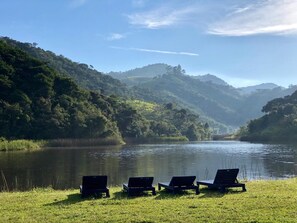 A serene lakeside view with three lounge chairs facing the water, surrounded by lush green hills. - FAZENDA CATUÇABA (São Luiz do Paraitinga)
