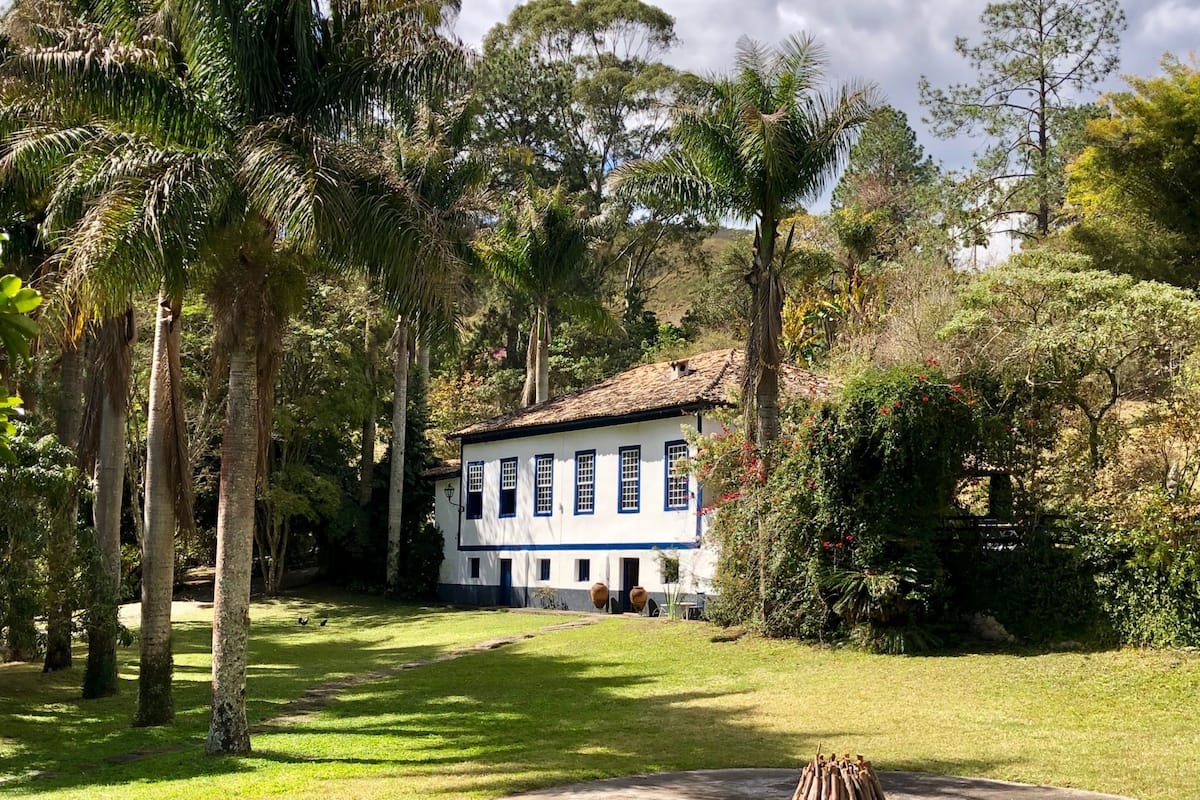 A white building with blue trim and shutters, surrounded by lush greenery and tall trees.