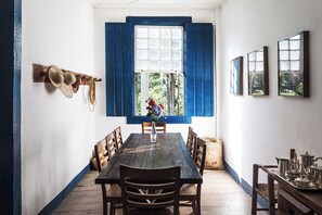A dining area with a wooden table, chairs, and a window with blue shutters. - FAZENDA CATUÇABA (São Luiz do Paraitinga)