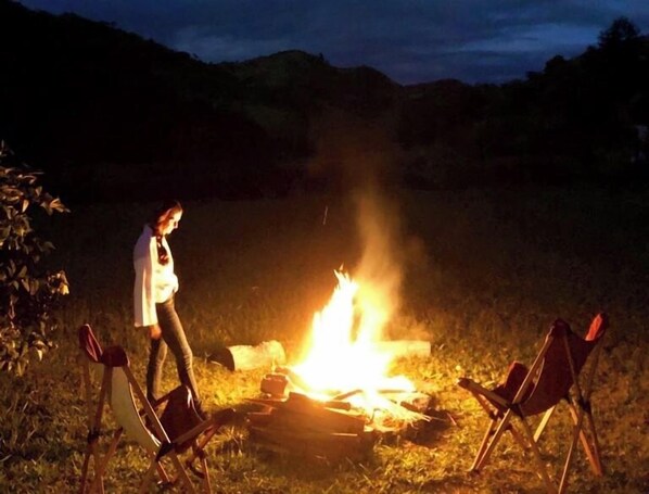 A person standing by a campfire with two chairs. - FAZENDA CATUÇABA (São Luiz do Paraitinga)