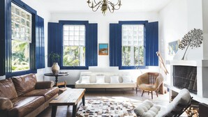 Ein Wohnzimmer mit einer braunen Ledersofa, einem weißen Sofa, einem gemusterten Teppich und einem weißen Sessel. Ein Kamin mit einer dekorativen Pflanze, eine Chandelier und ein großes Fenster mit blauen Fensterläden.