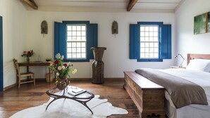 Ein Zimmer mit Schreibtisch aus Holz, einer großen Vase mit Blumen, einem Bett mit Holz-Kopfteil und zwei Fenstern mit blauen Fensterläden.