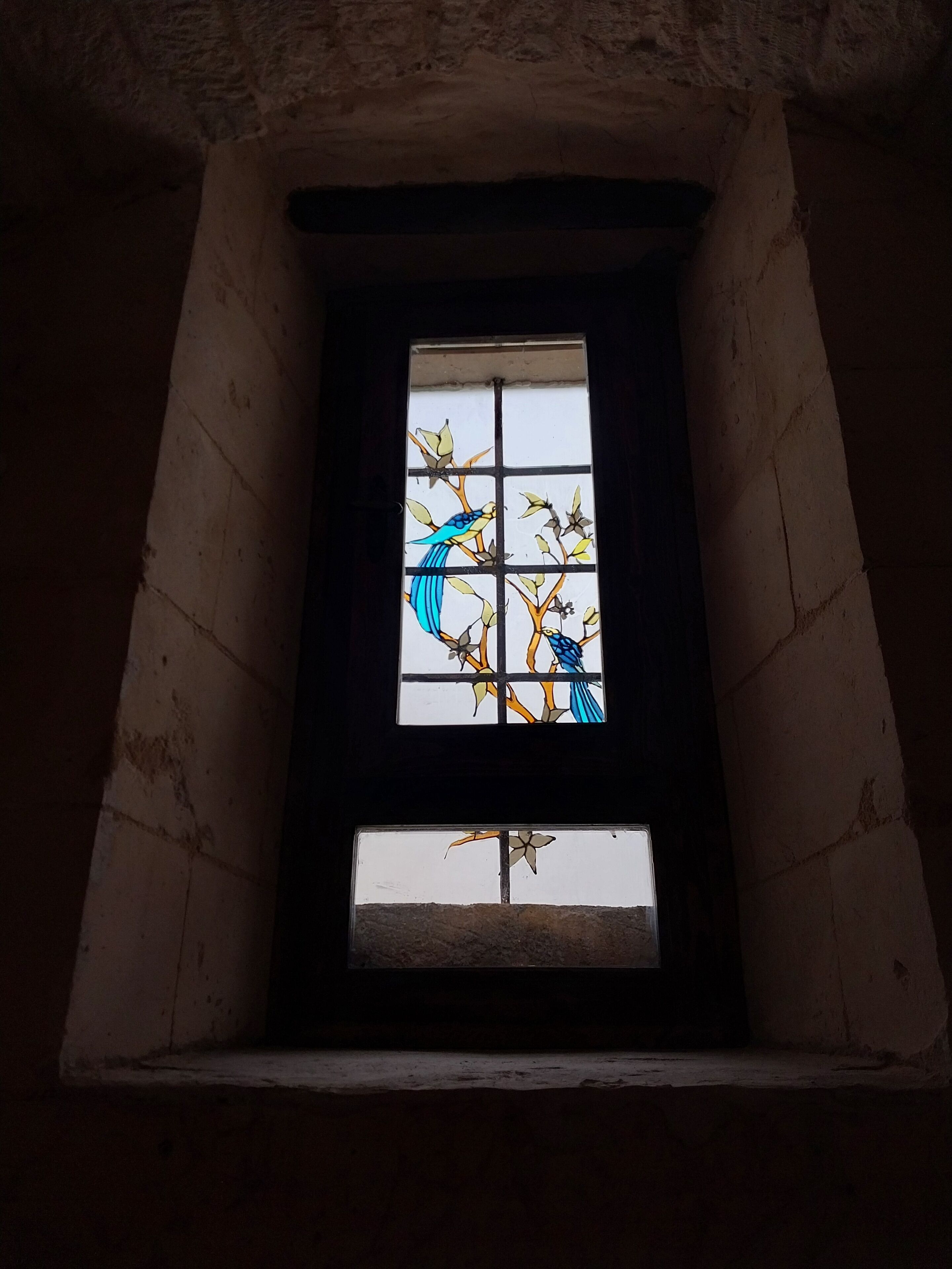 A stained glass window with a bird and floral design in a narrow, dimly lit room with stone walls.