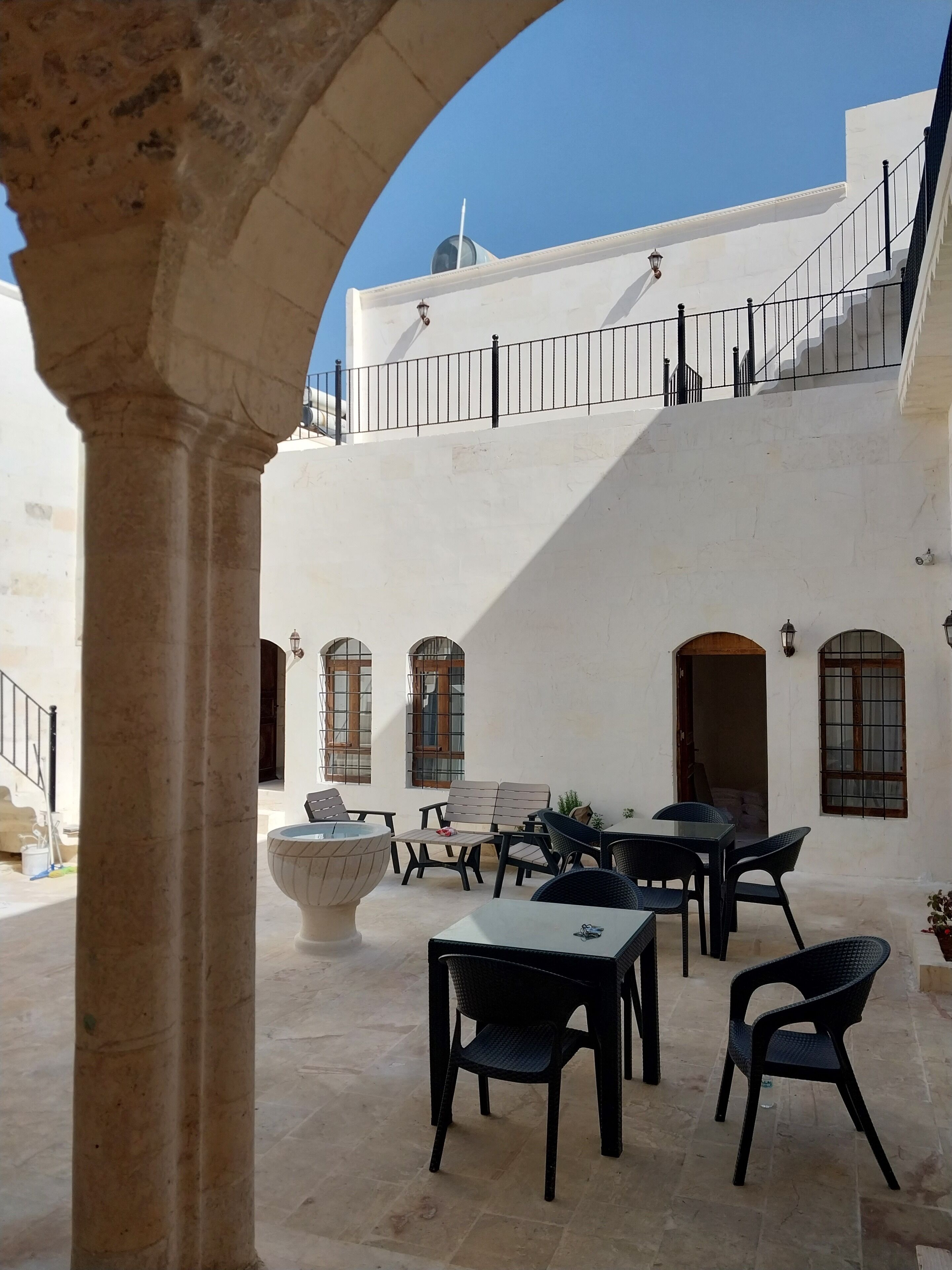 Un patio al aire libre con suelo de piedra, varias mesas y sillas, una fuente decorativa grande y una escalera que lleva a un nivel superior.