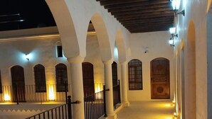 Un pasillo con columnas y arcos blancos que conduce a una puerta de madera con un reloj.