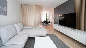 Ein modernes Wohnzimmer mit einem Ecksofa, einem Flachbildfernseher und einem Essbereich im Hintergrund.