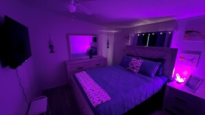 Une chambre à coucher comprenant un lit, une table de chevet, une commode, un ventilateur de plafond et un téléviseur fixé au mur.