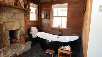 Une salle de bain rustique avec une baignoire sur pieds, une cheminée en pierre, des étagères en bois et un petit tabouret en bois.