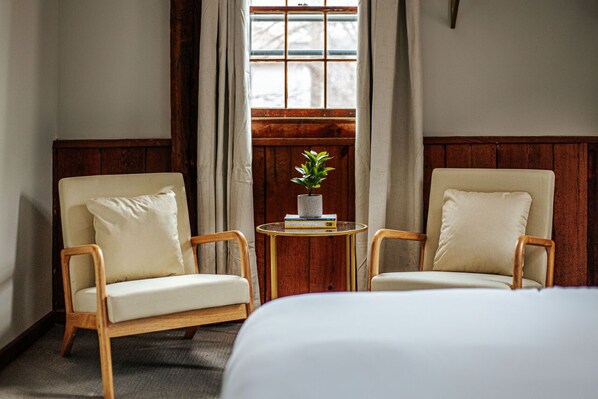 Une chambre d’hôtel avec deux fauteuils, une petite table ronde ornée d’une plante et de livres, et un grand lit.