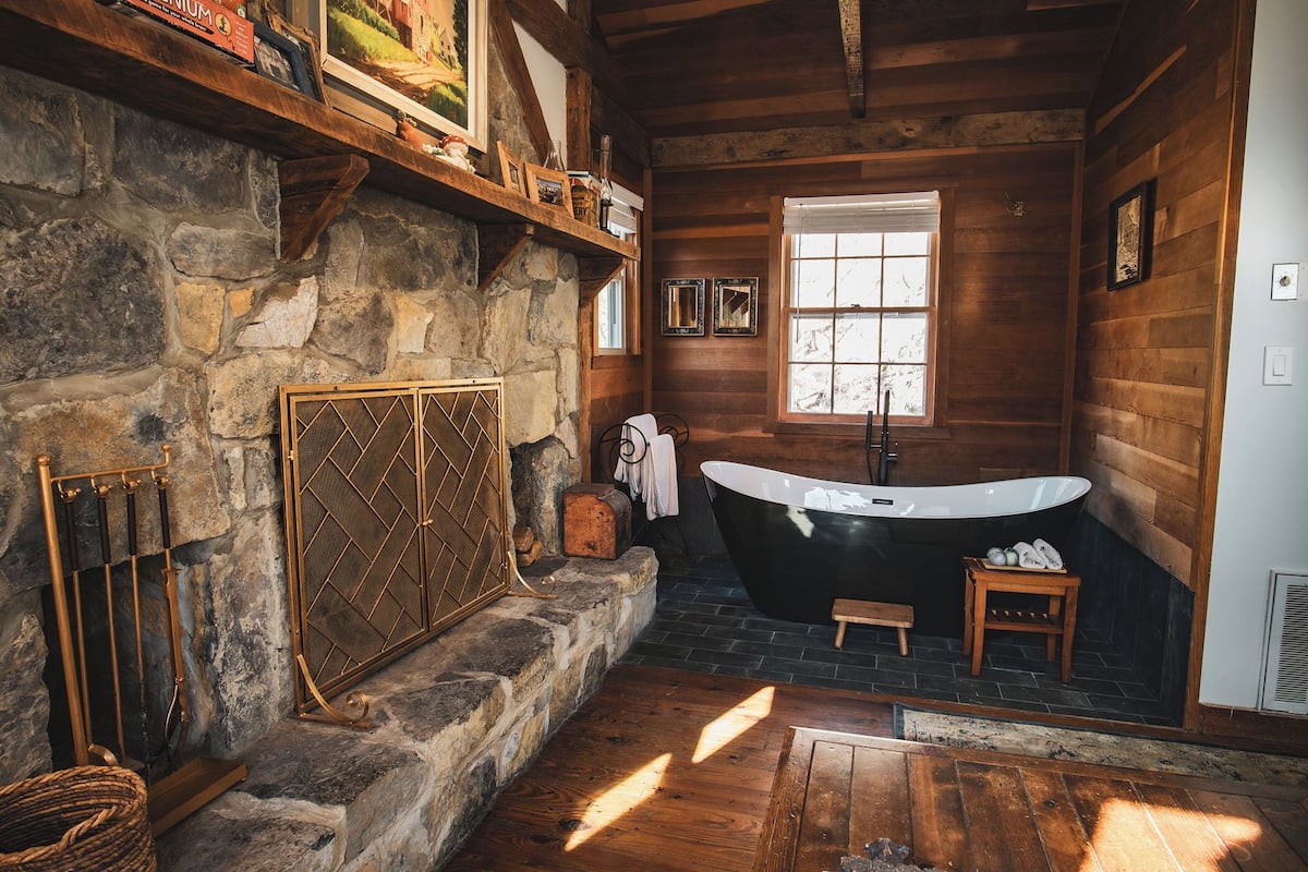 A rustic bathroom with a stone wall, wooden beams, a freestanding bathtub, and a window providing natural light.