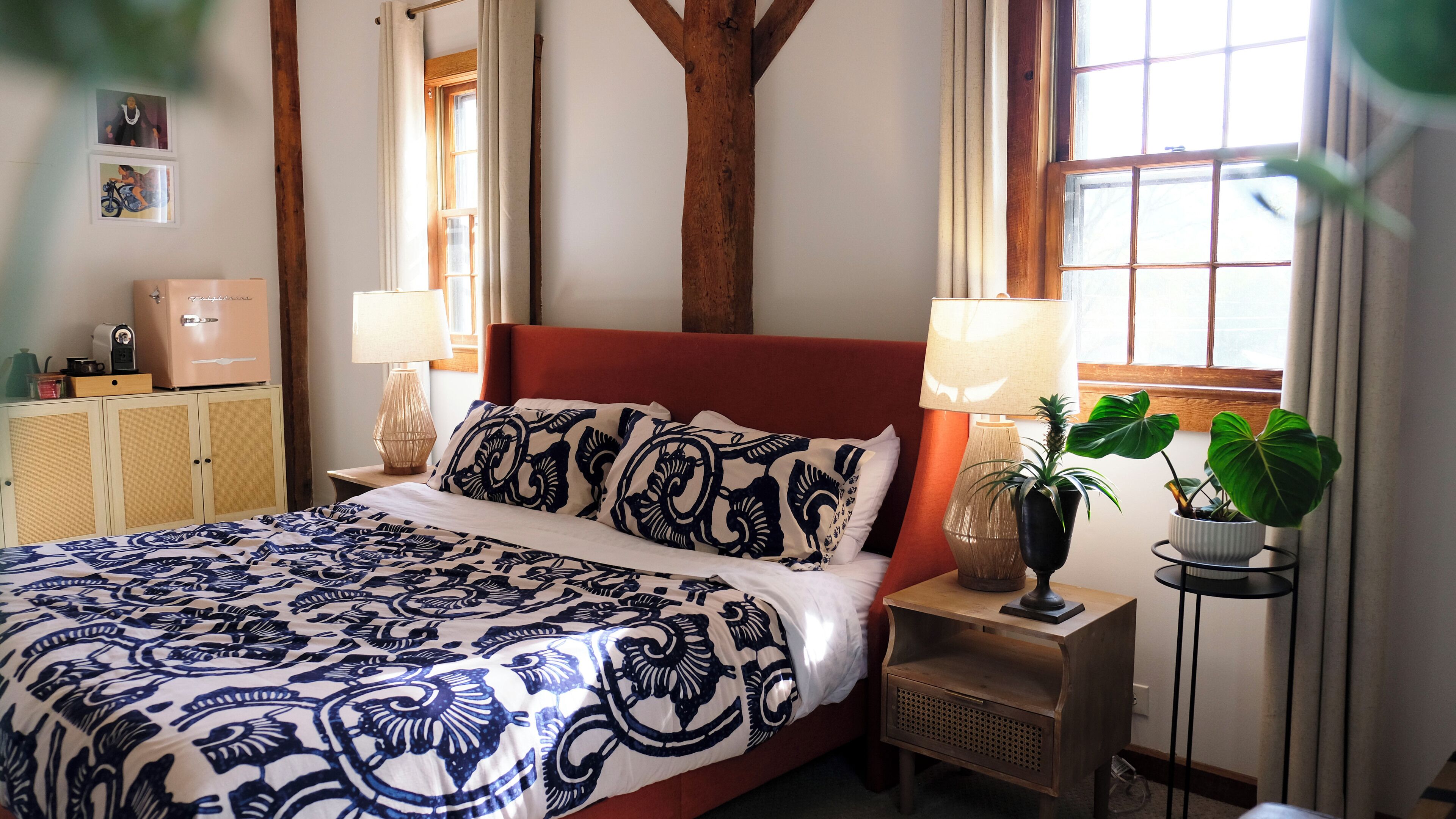 Cama bem arrumada com roupa de cama estampada, cabeceira de madeira, abajures, uma pequena mesa com uma planta e um guarda-roupa com espelho e máquina de café.