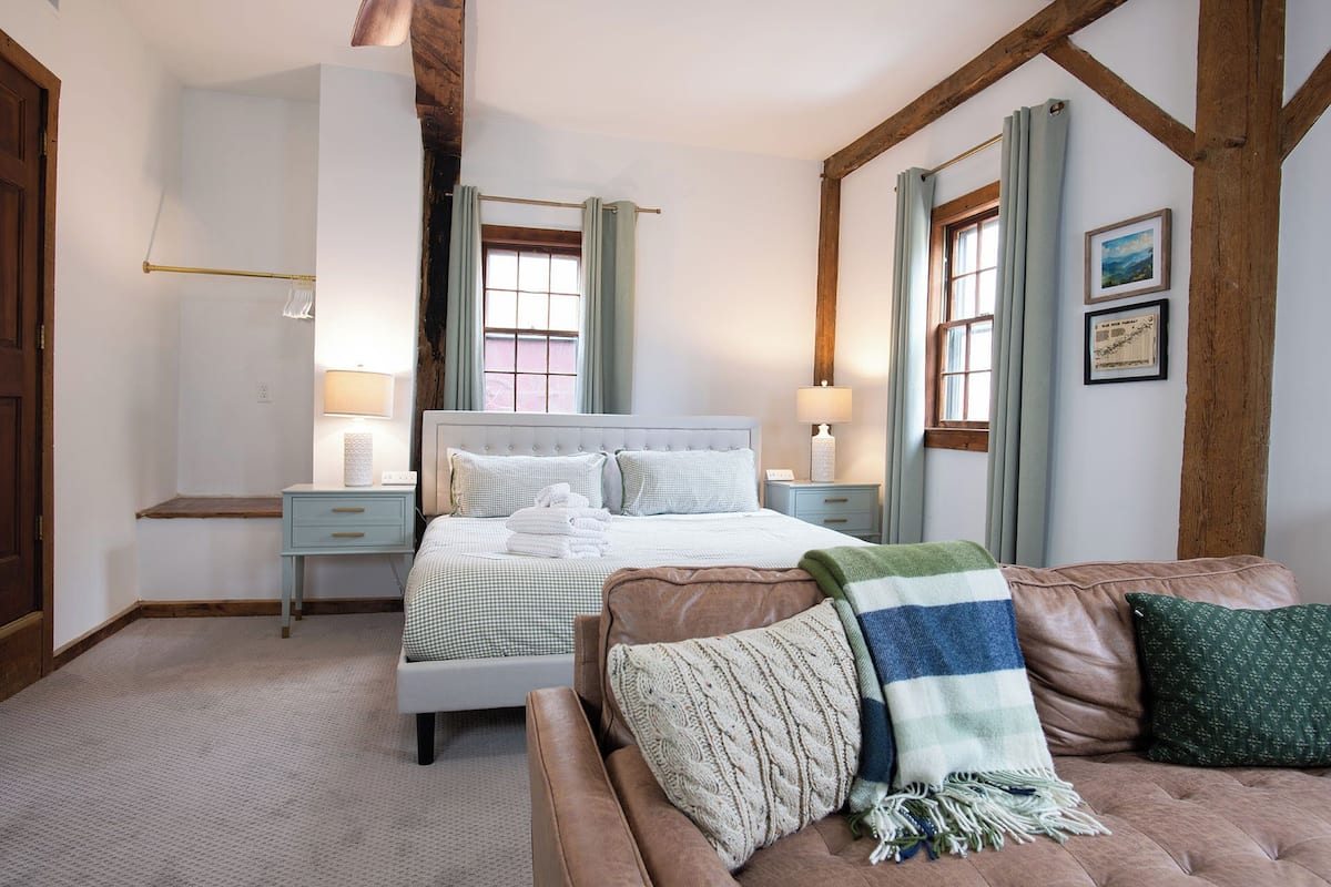 A bedroom with a bed, a nightstand, a dresser, a window with curtains, and a wooden ceiling beam.