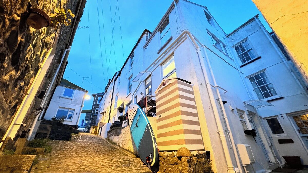 A narrow cobblestone street flanked by white buildings with a blue surfboard leaning against one wall.