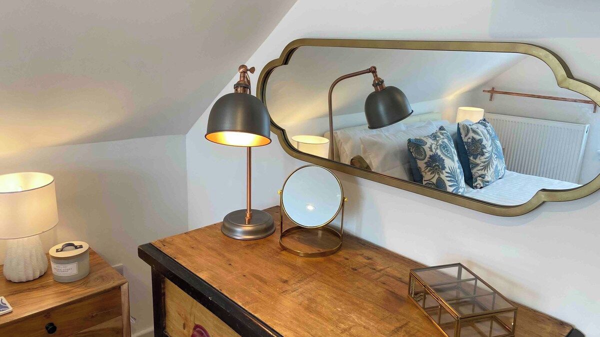 Een slaapkamer met een houten dressoir, een spiegel, twee lampen, en een nachtkastje met een lamp en een doos.