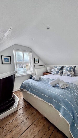 Een slaapkamer met een schuine plafond, een bed met een deken met blauw-wit patroon, een radiator, een raam met zonwering, en een lijst met een afbeelding aan de muur.