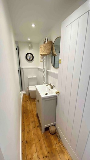 A narrow hallway with a wooden floor, a white sink with a gold faucet, a white toilet, and a white door.