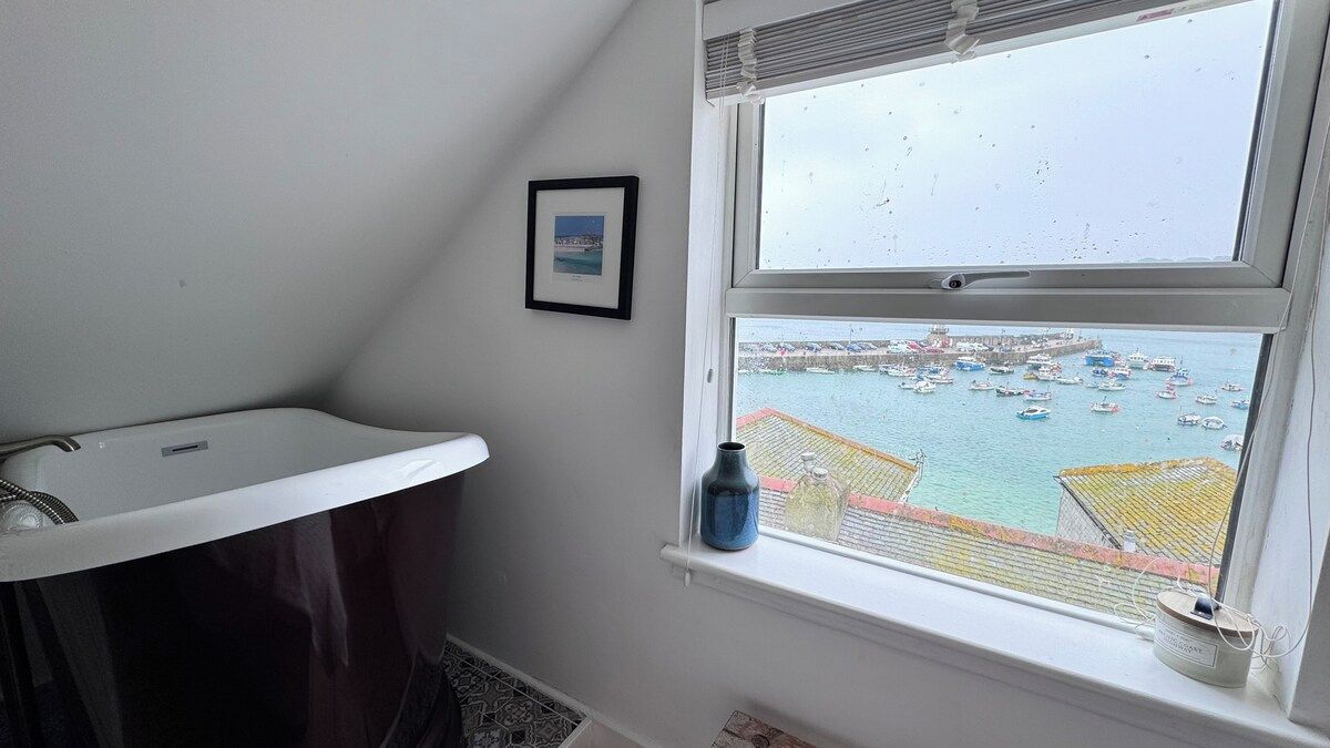 Casa de banho com lavatório branco, um quadro na parede e uma janela com vista para uma marina e barcos.