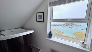 Une salle de bain avec un lavabo blanc, un tableau encadré au mur et une fenêtre donnant sur un port où sont amarrés des bateaux.