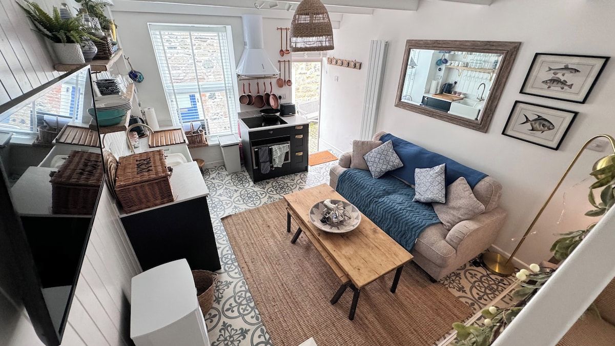 A modern kitchen with a stove, sink, and hanging pots. There is a dining area with a wooden table and a sofa.