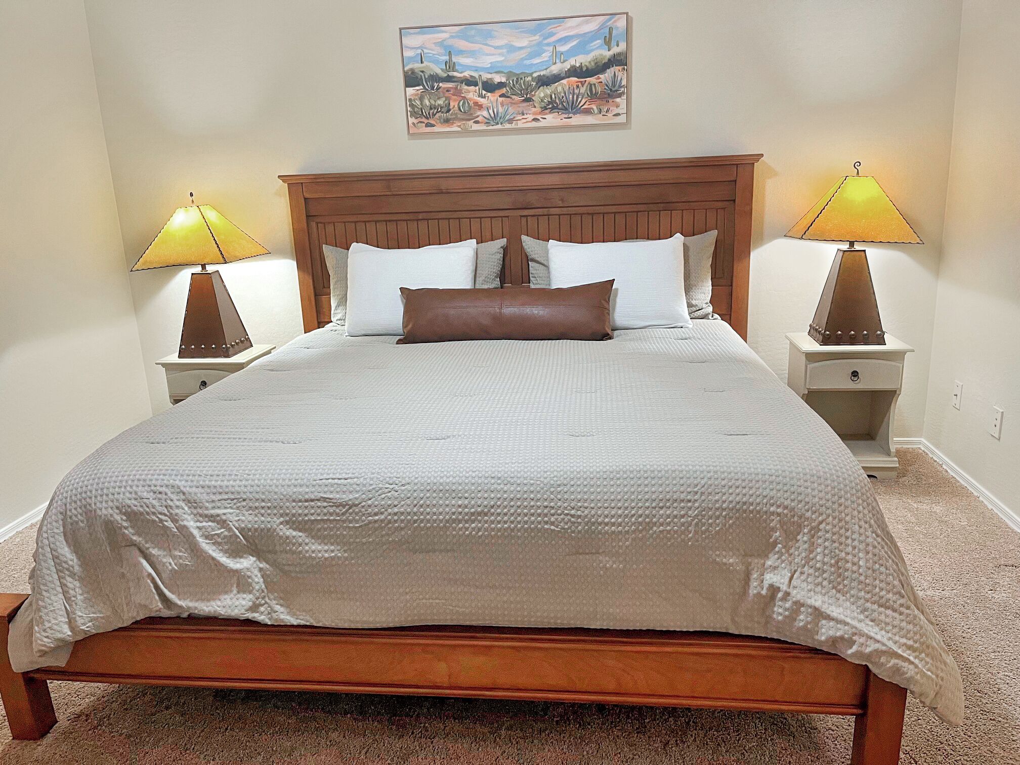 A neatly made bed with a wooden headboard, two bedside tables with lamps, and a painting on the wall.